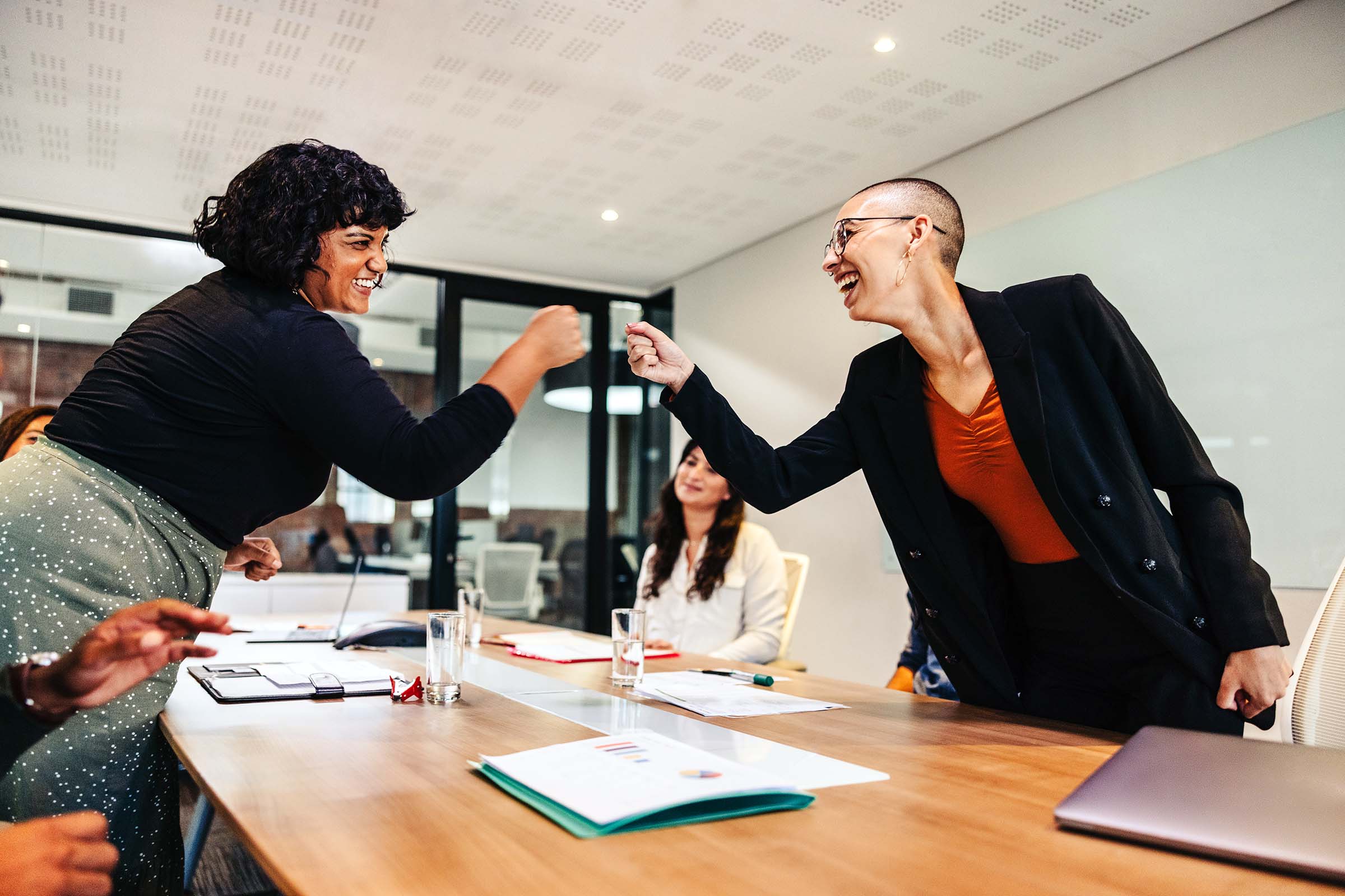 Two business professionals smiling and fist-bumping across a conference table during a meeting, with a third person observing in the background.