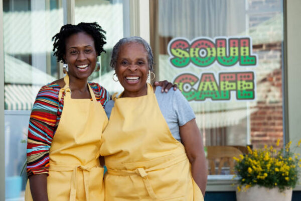 Two women, one younger and one older standing in front of their business smiling with yellow aprons on.