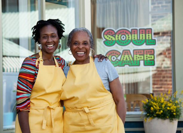 Two women, one younger and one older standing in front of their business smiling with yellow aprons on.