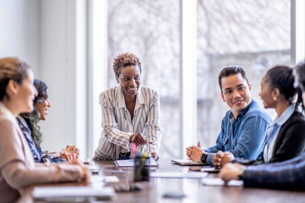 A professional woman standing and leading a discussion at a conference table, with colleagues seated and listening attentively