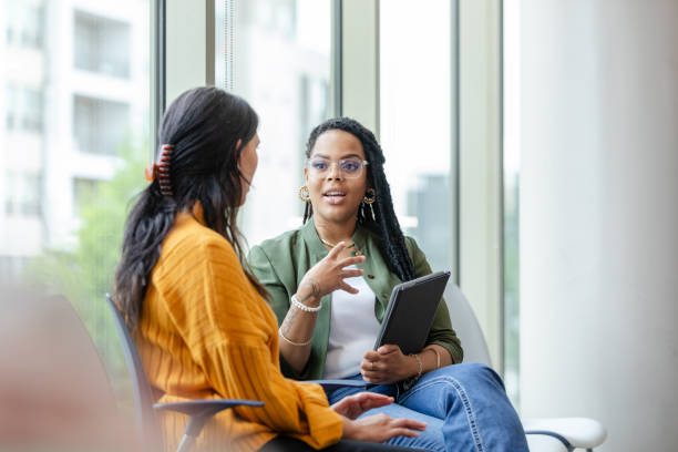 Two women having a professional conversation in a bright office space, with one holding a tablet and actively engaging the other.