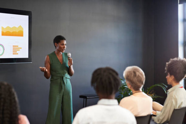 Woman in green jumpsuit, standing at the front of the room speaking to a group of four people