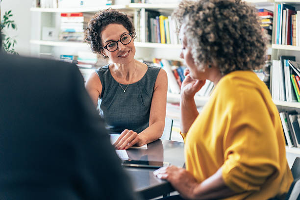 Three professionals engaged in a friendly business discussion in an office setting, with one woman smiling and actively listening across the table.