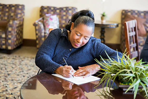 Woman sitting at a table in a comfortable lounge area, focused on filling out paperwork or a form with a pen.