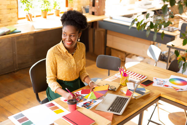Smiling woman working in a bright studio space, surrounded by color swatches, design tools, and a laptop, representing creativity and entrepreneurship