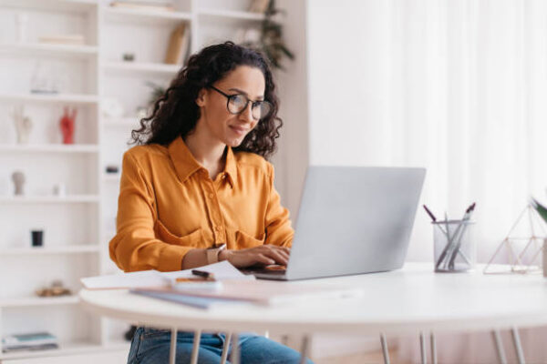 Woman wearing glasses and an orange blouse working on a laptop at a bright, modern desk. Papers and office supplies are on the table around her, and shelves are visible in the background.