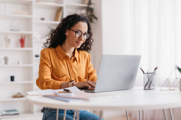 Woman wearing glasses and an orange blouse working on a laptop at a bright, modern desk. Papers and office supplies are on the table around her, and shelves are visible in the background.