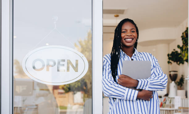 Woman smiling in front of store window, sign says open. She is wearing a blue stripped shirt and holding a laptop
