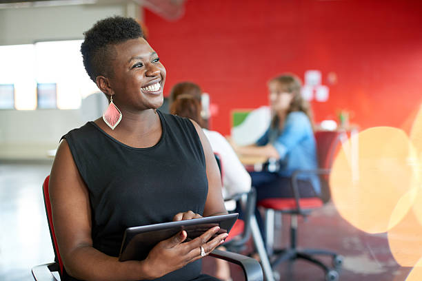 Smiling Black woman holding a tablet in a bright coworking space, with a collaborative team meeting in the background
