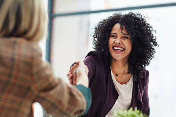 Two women connecting in a professional setting, smiling and shaking hands during a meeting.