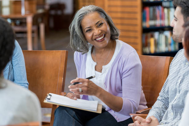 Smiling older woman holding a clipboard and pen, leading a group discussion in a library or classroom setting with others seated around her.