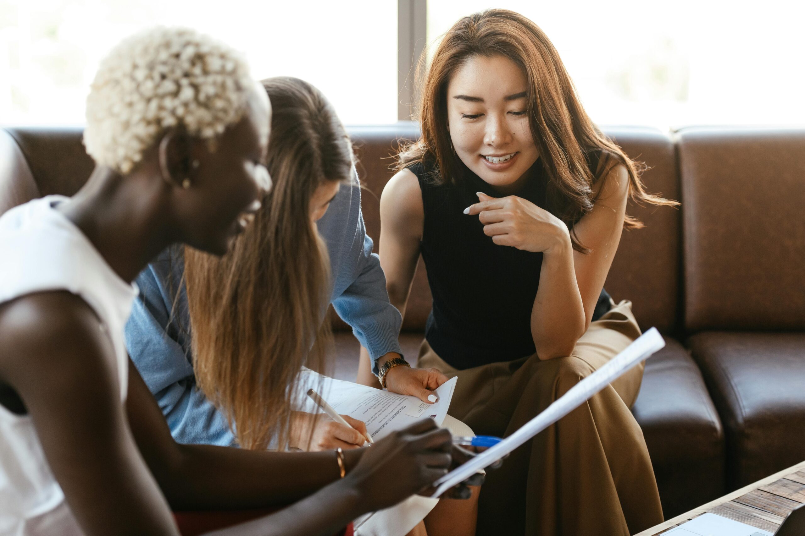 Three women sit together at a table, smiling and working on paperwork or notes.
