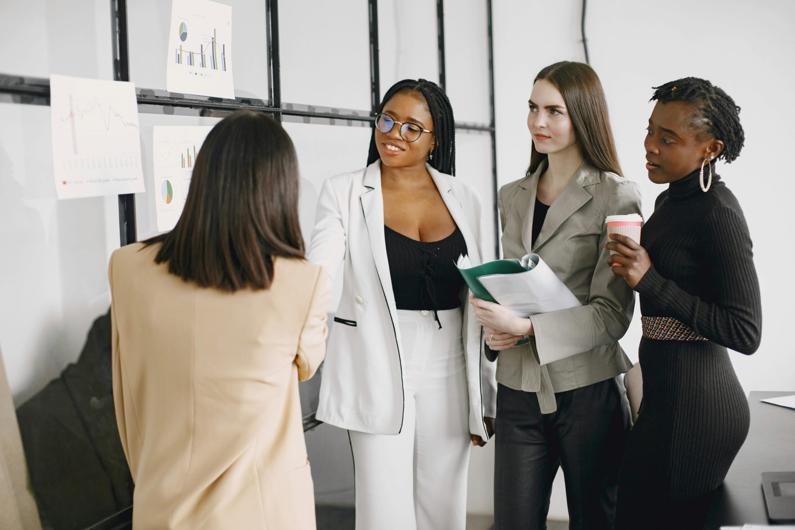 Four professional women engaged in a collaborative discussion in front of data charts, sharing ideas and strategies in a modern office setting