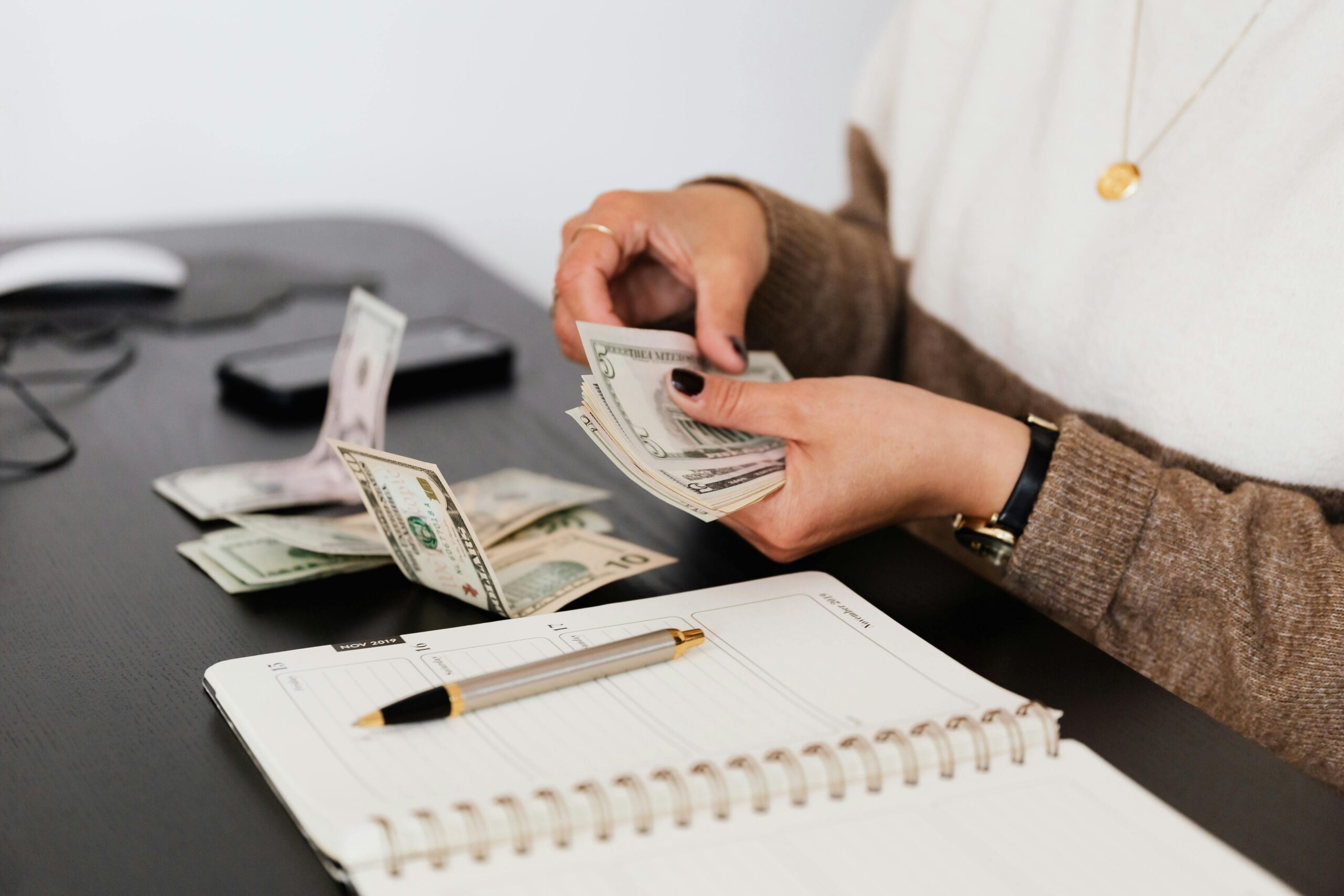 Person counting cash at a desk with a notebook and pen, preparing finances or budgeting for business expenses.