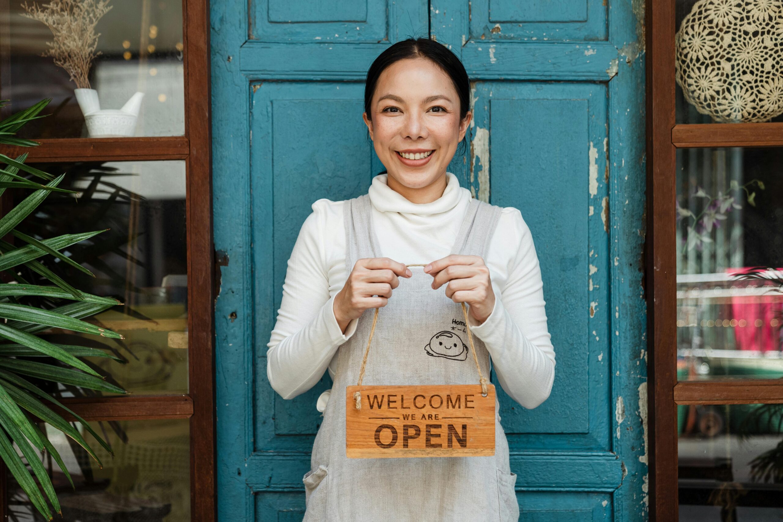 Smiling small business owner standing in front of a shop door, holding a wooden 'Welcome, We Are Open' sign.