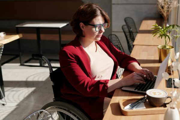 Woman using a wheelchair working on a laptop at a sunlit café table, focused on her tasks.