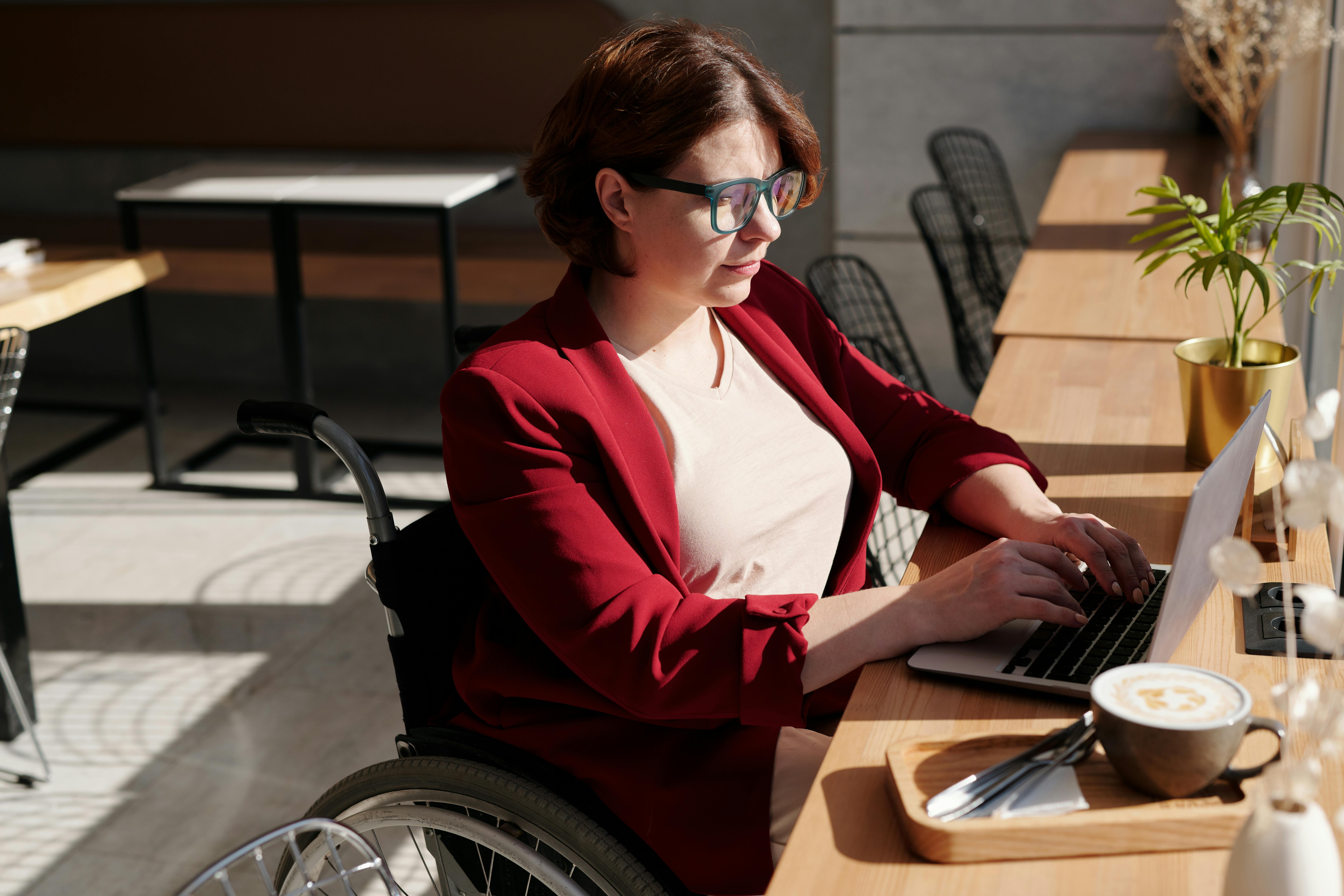 Woman using a wheelchair working on a laptop at a sunlit café table, focused on her tasks.