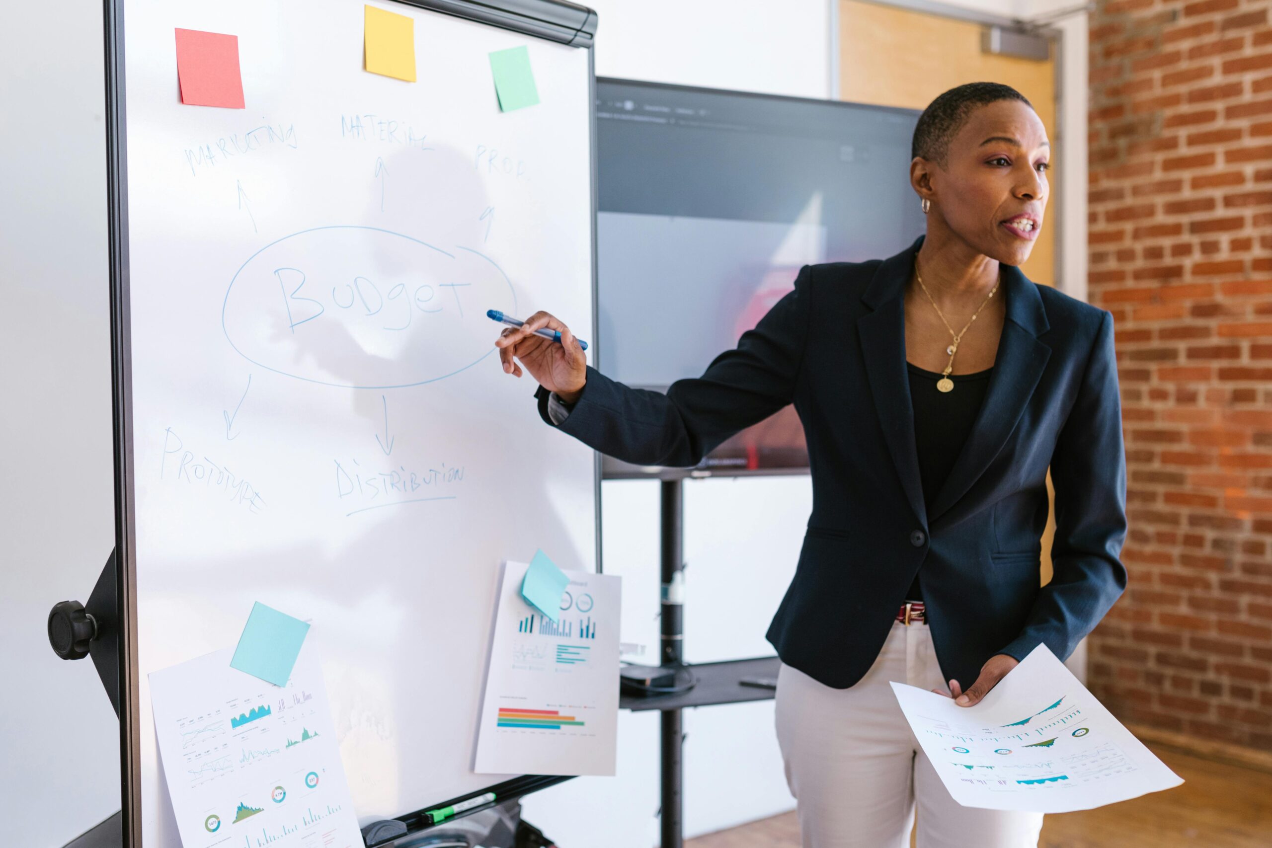 Professional woman giving a business presentation on budgeting, pointing to a whiteboard with charts and strategy notes during a workshop or training session.