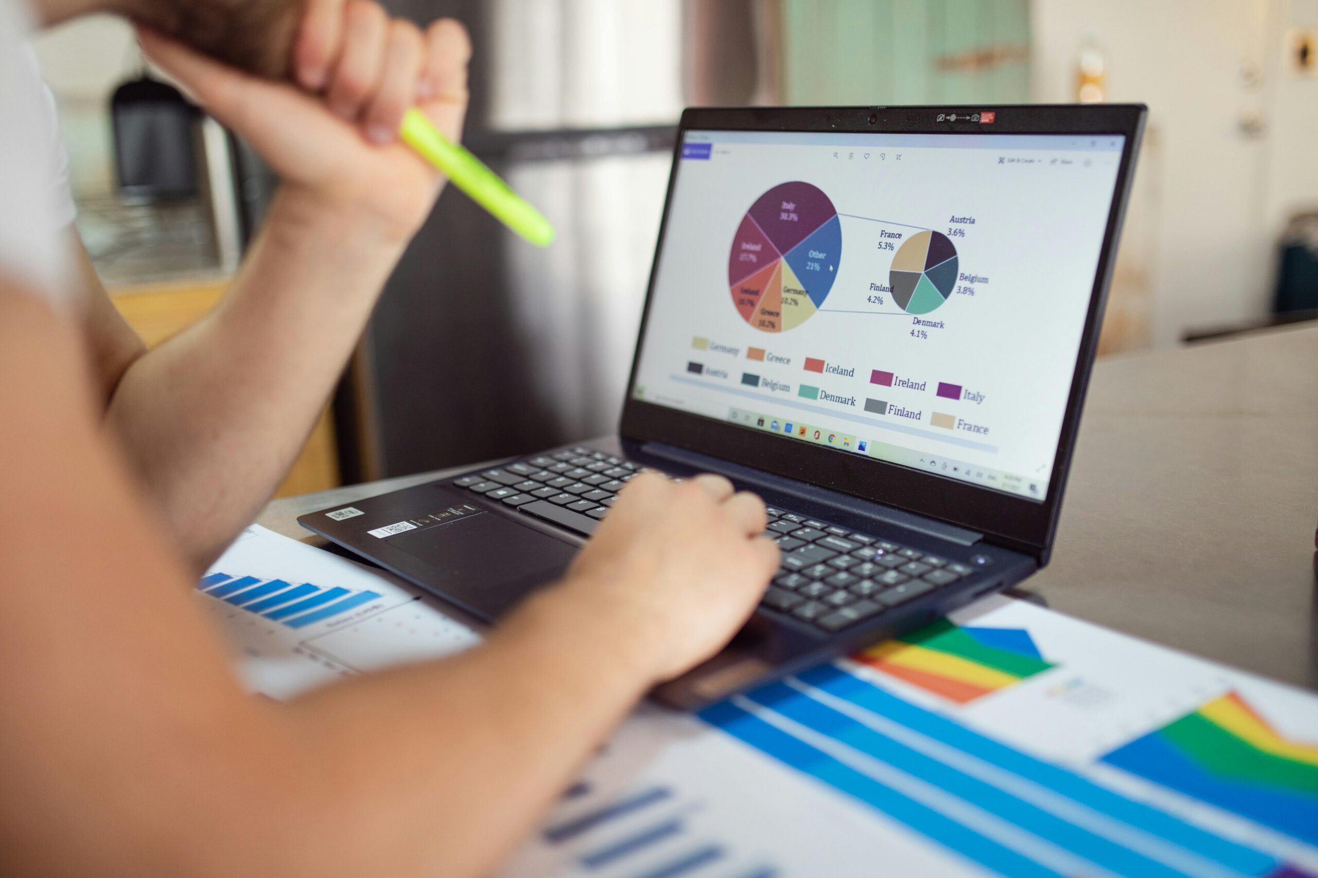 Person analyzing colorful financial charts and graphs on a laptop, with printed data reports spread out on a desk