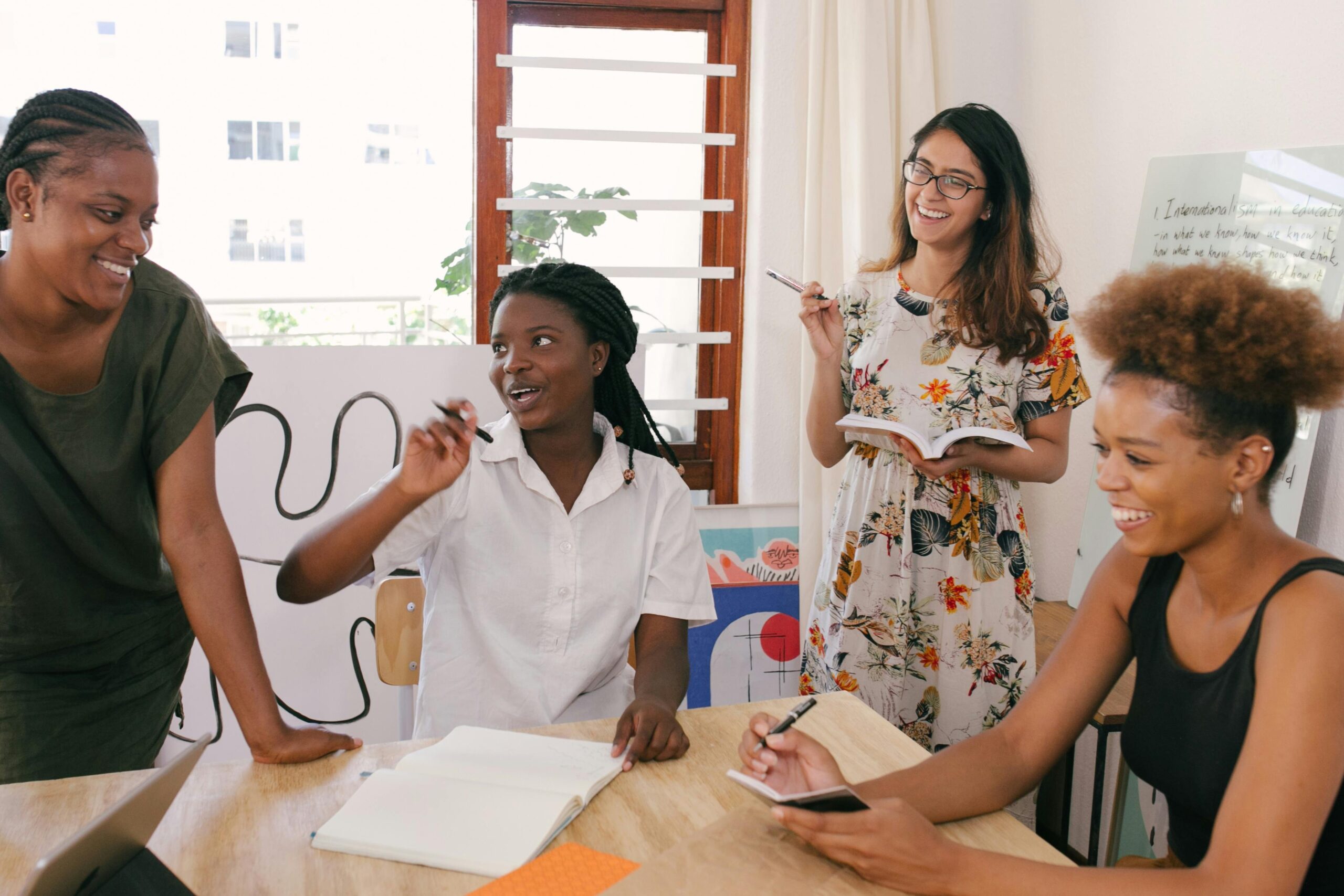 Four women gathered around a table, smiling and collaborating during a casual brainstorming session in a bright, creative workspace